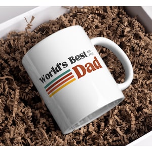 May include: White ceramic mug with the text "World's Best Dad" in black, red, and retro stripes. The mug has a curved handle and is presented in a gift box with brown paper filler.