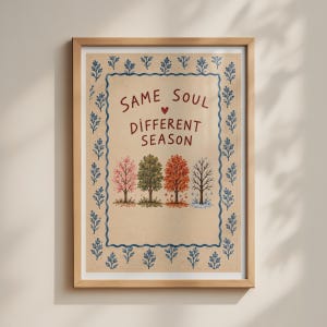 May include: A framed wall art print on a beige background featuring the text SAME SOUL DIFFERENT SEASON with a small red heart. Below the text are four trees representing different seasons, surrounded by a blue floral border pattern.
