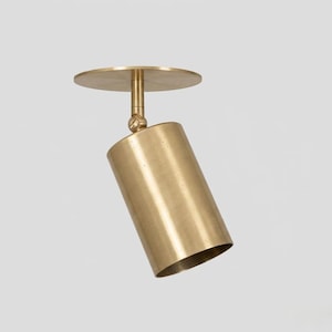 May include: A brass ceiling spotlight featuring a circular mounting plate and a cylindrical head attached by a swivel joint. The metal surface has a brushed finish, and the fixture is designed to be mounted directly to a flat surface for directional lighting.