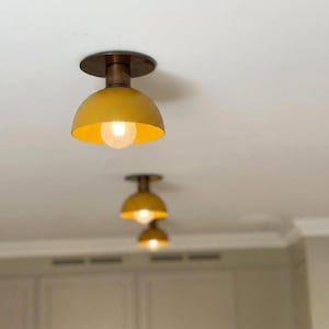 Może przedstawiać: Zbliżenie trzech żółtych lamp sufitowych w kształcie kopuły z odsłoniętymi żarówkami. Lampy mają podstawę w kolorze mosiądzu i są zamontowane na białym suficie. Światła są ustawione w rzędzie, z najbliższym w ostrości.