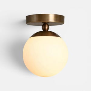 May include: A modern ceiling light fixture with a round, frosted glass globe. The fixture has a bronze-colored metal base and a small, spherical detail connecting the globe to the ceiling mount. The light is designed to be mounted on a ceiling.