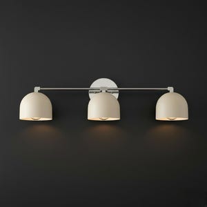 May include: A three-light vanity wall fixture featuring a horizontal brushed nickel bar and a circular wall mount. Three matte cream-coloured dome-shaped shades are evenly spaced along the bar, each housing a single light bulb pointing downward.