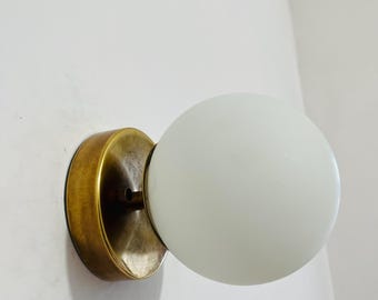 May include: A wall-mounted light fixture with a round, frosted white glass globe. The fixture has a circular, antique brass-colored base. The light is mounted on a white wall.
