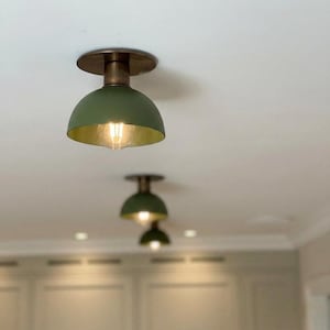 Könnte beinhalten: Olivgrüne, kuppelförmige Deckenleuchten mit goldfarbenem Innenraum und Messingbeschlägen. Die Leuchten sind an einer weißen Decke montiert, wobei zwei weitere Leuchten im Hintergrund sichtbar sind. Die Lichter sind eingeschaltet und beleuchten den Raum.