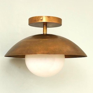 Pode incluir: Uma luminária de teto semi-embutida com uma cúpula larga em forma de domo com acabamento em latão antigo escovado. Um globo esférico de vidro branco opaco está centralizado sob o domo de metal, fixado a uma canopla circular correspondente.