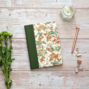 May include: A green and white journal with a floral pattern. The journal has a green spine and a white cover with a floral pattern in shades of red, green, and white. The journal is sitting on a white wooden surface.