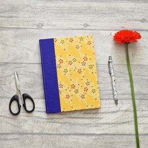 May include: A yellow notebook with a blue spine and a floral pattern. The notebook is on a light wood surface with a red flower and a pen.
