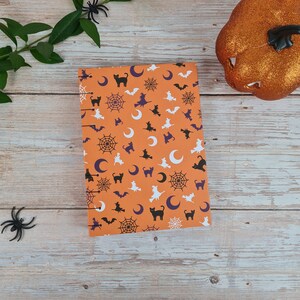 Op de afbeelding: Oranje notitieboek met een Halloween-thema. De omslag heeft een herhalend patroon van zwarte katten, vleermuizen, halve manen, spinnen en spinnenwebben op een oranje achtergrond.