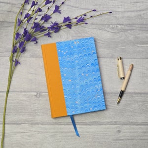 May include: A blue and orange journal with a marbled cover and a blue ribbon bookmark. A gold pen is lying on a wooden surface next to the journal.