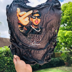 May include: A black t-shirt featuring a graphic print of a person wearing sunglasses and a cap, making a hand gesture. The text BREEZY BOWL XX is printed below the image, accompanied by a signature. The fabric has a gathered, textured appearance.