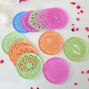 May include: A collection of colorful, round coasters in shades of blue, pink, green, and orange. Each coaster features intricate, cut-out designs, including floral patterns and other decorative motifs. The coasters are arranged on a white surface.