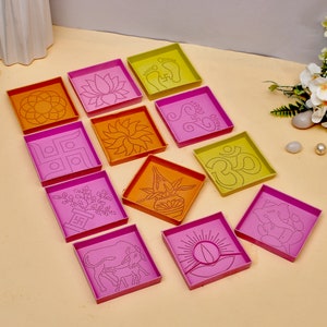 Könnte beinhalten: Satz von zehn quadratischen Schablonen mit verschiedenen Designs, darunter eine Lotusblume, ein Swastika, eine Sonne und eine Diya. Die Schablonen sind aus rosa und orangefarbenem Kunststoff.