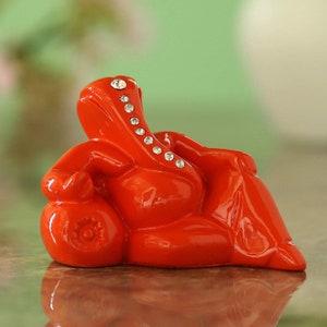 Könnte beinhalten: Eine rote, glänzende Keramikfigur von Ganesha, dem hinduistischen Gott der Weisheit und des Wohlstands, der mit gekreuztem Bein und der Hand auf dem Knie sitzt. Die Figur ist mit Strasssteinen verziert.