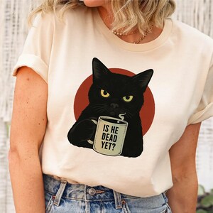 May include: Cream-colored t-shirt featuring a black cat illustration holding a mug with the text "IS HE DEAD YET?" The cat is set against a red circle. The shirt is a casual wear item.