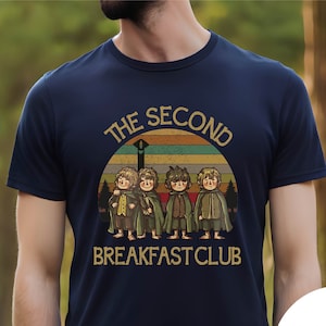 May include: Navy blue t-shirt featuring a retro design with the text "THE SECOND BREAKFAST CLUB" above an illustration of four hobbits. The design is set against a sunset-colored background. The shirt is a classic crew neck.