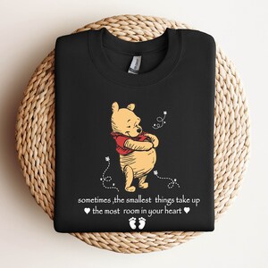 Peut inclure: T-shirt noir avec une illustration de Winnie l'ourson et le texte "sometimes, the smallest things take up the most room in your heart".