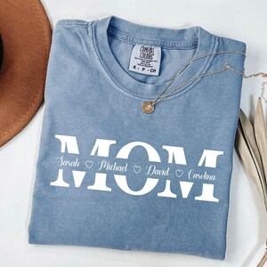 May include: A light blue t-shirt with the word "MOM" in white letters. The names "Sarah", "Michael", "David", and "Carolina" are written below the letters with hearts between each name.