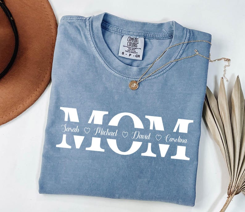 May include: A light blue t-shirt with the word "MOM" in white letters. The names "Sarah", "Michael", "David", and "Carolina" are written below the letters with hearts between each name.