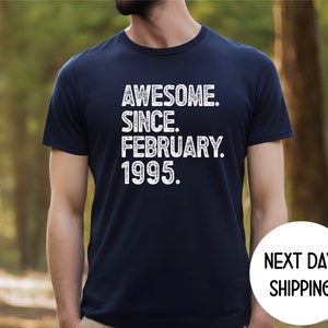 May include: Navy blue t-shirt with the text "AWESOME. SINCE. FEBRUARY. 1995." in white. The shirt has a classic crew neck and short sleeves. The words are stacked vertically. The shirt is worn by a person.