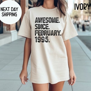 May include: Ivory-colored t-shirt with the text "AWESOME. SINCE. FEBRUARY. 1995." in black. The shirt has a crew neck and short sleeves. The words "NEXT DAY SHIPPING" are in a white circle in the upper left corner.