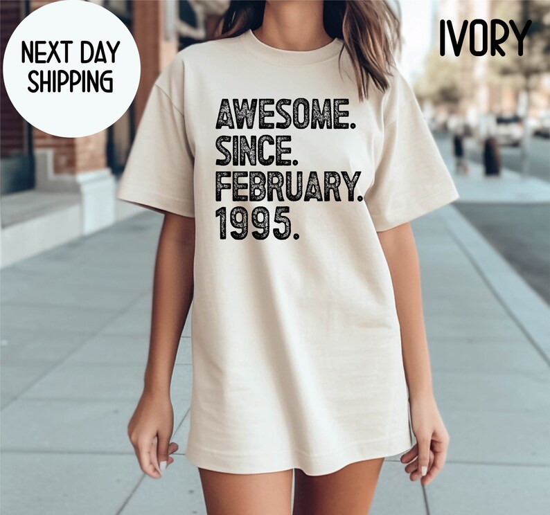 May include: Ivory-colored t-shirt with the text "AWESOME. SINCE. FEBRUARY. 1995." in black. The shirt has a crew neck and short sleeves. The words "NEXT DAY SHIPPING" are in a white circle in the upper left corner.