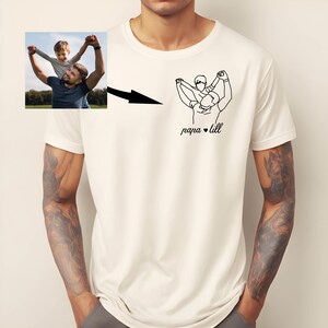May include: Cream-colored t-shirt featuring a black line drawing of a person carrying a child on their shoulders, with the text "papa &bull; till" below. A small photo of a father and child is in the upper left corner.