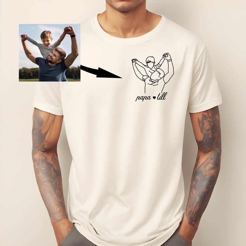 May include: Cream-colored t-shirt featuring a black line drawing of a person carrying a child on their shoulders, with the text "papa &bull; till" below. A small photo of a father and child is in the upper left corner.