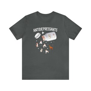 May include: Dark gray t-shirt featuring a graphic of a pill bottle labeled "ANTIDEPRESSANTS" with cartoon dogs spilling out. The dogs are various breeds and colors, with hearts scattered around the design. A fun, lighthearted design.