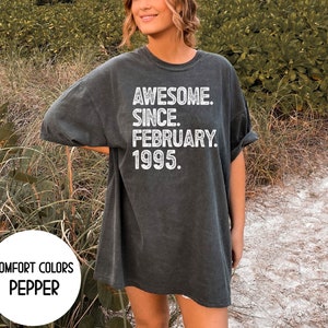 May include: A dark gray t-shirt with the text "AWESOME. SINCE. FEBRUARY. 1995." in white. The shirt also has the words "COMFORT COLORS" and "PEPPER" in white. The shirt is a relaxed fit.