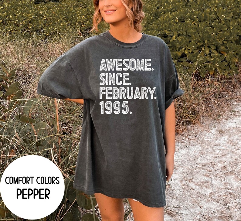 May include: A dark gray t-shirt with the text "AWESOME. SINCE. FEBRUARY. 1995." in white. The shirt also has the words "COMFORT COLORS" and "PEPPER" in white. The shirt is a relaxed fit.