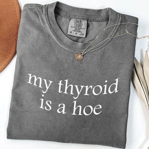 May include: A gray Comfort Colors t-shirt with the phrase "my thyroid is a hoe" in white lettering. A gold-colored necklace with a pendant rests on the shirt. The shirt is folded and has a small tag.