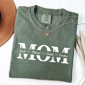 May include: A green t-shirt with the word "MOM" in white letters. The names "Sarah", "Michael", "David", and "Carolina" are written below the letters with hearts between each name.