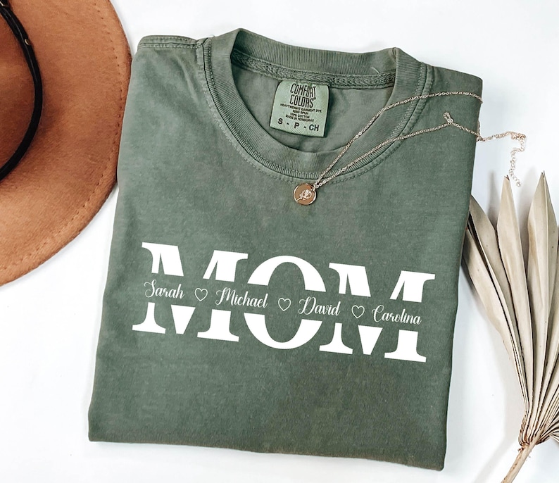 May include: A green t-shirt with the word "MOM" in white letters. The names "Sarah", "Michael", "David", and "Carolina" are written below the letters with hearts between each name.