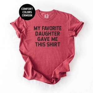 May include: Crimson red t-shirt with rolled sleeves. The shirt features the text "MY FAVORITE DAUGHTER GAVE ME THIS SHIRT" in black block letters. A black circle in the upper left corner reads "COMFORT COLORS CRIMSON".