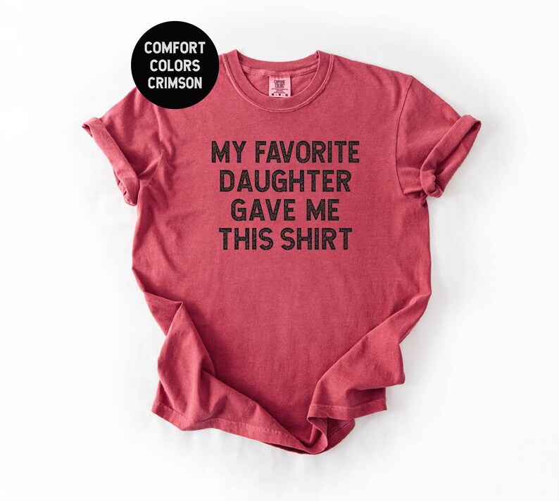 May include: Crimson red t-shirt with rolled sleeves. The shirt features the text "MY FAVORITE DAUGHTER GAVE ME THIS SHIRT" in black block letters. A black circle in the upper left corner reads "COMFORT COLORS CRIMSON".