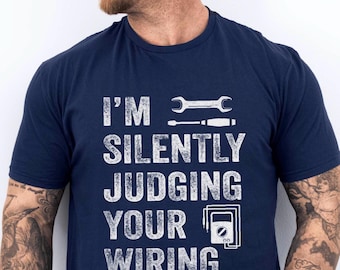 May include: Navy blue t-shirt with the text "I'M SILENTLY JUDGING YOUR WIRING" in white. The design includes a wrench, screwdriver, and electrical meter. A humorous shirt for electricians or those in the electrical trade.