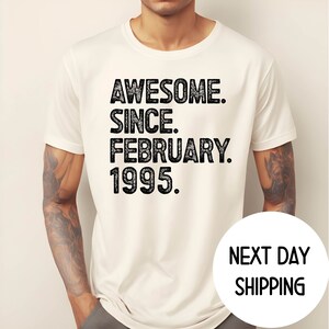 May include: Cream-colored t-shirt with the text "AWESOME. SINCE. FEBRUARY. 1995." in a distressed black font. The shirt is short-sleeved and appears to be made of a soft material. The shirt is being worn by a person.