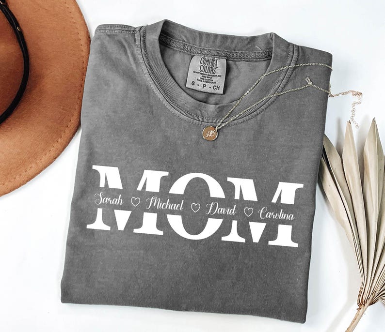 May include: Gray t-shirt with the word "MOM" in large white letters. The names "Sarah", "Michael", "David", and "Carolina" are written in smaller white letters below the word "MOM" with a heart symbol between each name.