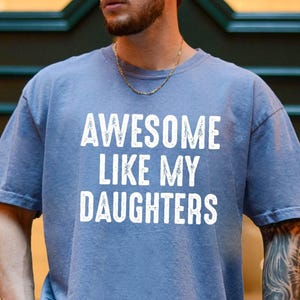 May include: A blue short-sleeved t-shirt with the text "AWESOME LIKE MY DAUGHTERS" in white block letters. The shirt has a crew neck and a relaxed fit. The person wearing the shirt has a gold chain.