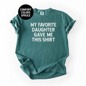 May include: Spruce-colored t-shirt with the text "MY FAVORITE DAUGHTER GAVE ME THIS SHIRT" in white block letters. The shirt is a crew neck style and has rolled-up sleeves. The Comfort Colors brand is visible.