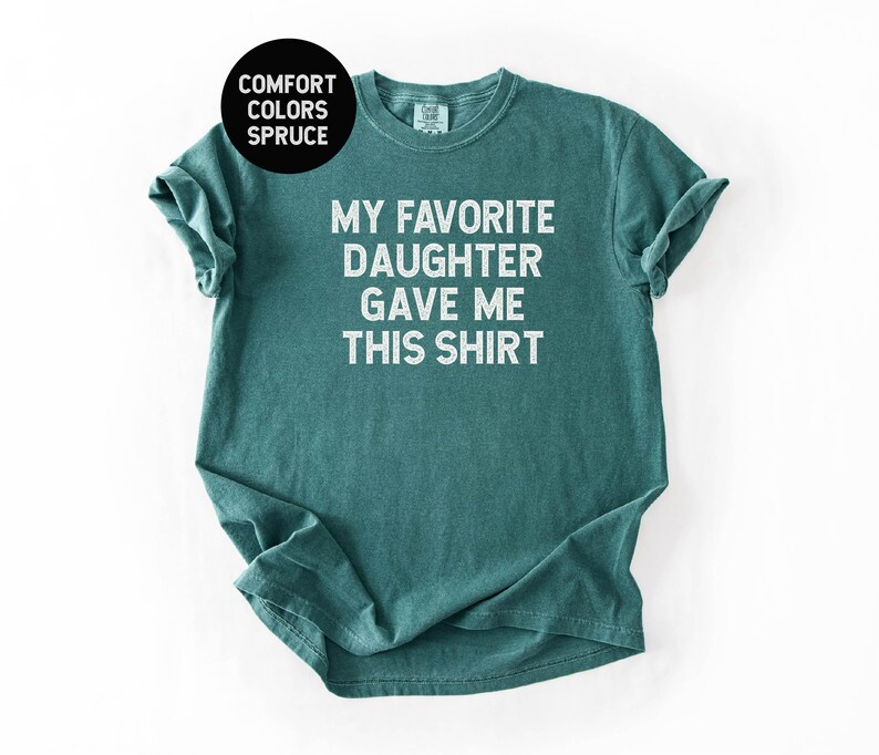 May include: Spruce-colored t-shirt with the text "MY FAVORITE DAUGHTER GAVE ME THIS SHIRT" in white block letters. The shirt is a crew neck style and has rolled-up sleeves. The Comfort Colors brand is visible.