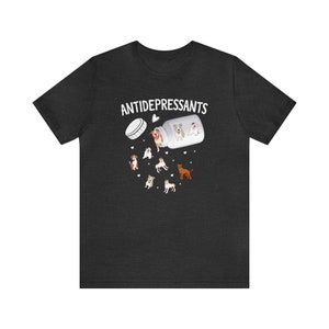May include: Dark gray t-shirt featuring a graphic of a pill bottle labeled "ANTIDEPRESSANTS" with cartoon dogs spilling out. The design includes various dog breeds and small heart shapes, creating a playful and whimsical aesthetic.