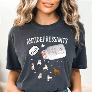 May include: Dark gray t-shirt with the word "ANTIDEPRESSANTS" in white. The shirt features a graphic of a pill bottle spilling out cartoon dogs. The dogs are in various poses and colors, with small white hearts scattered around.
