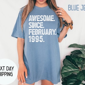 May include: A blue jean colored t-shirt with the text "AWESOME. SINCE. FEBRUARY. 1995." in white. The shirt is a casual style, with a relaxed fit. The words "BLUE JEAN" are printed in the upper right corner.