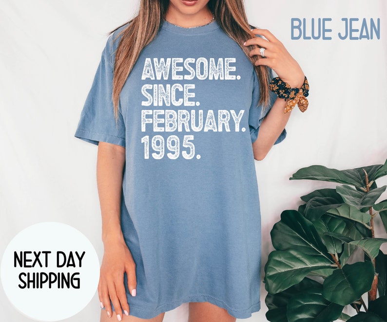 May include: A blue jean colored t-shirt with the text "AWESOME. SINCE. FEBRUARY. 1995." in white. The shirt is a casual style, with a relaxed fit. The words "BLUE JEAN" are printed in the upper right corner.