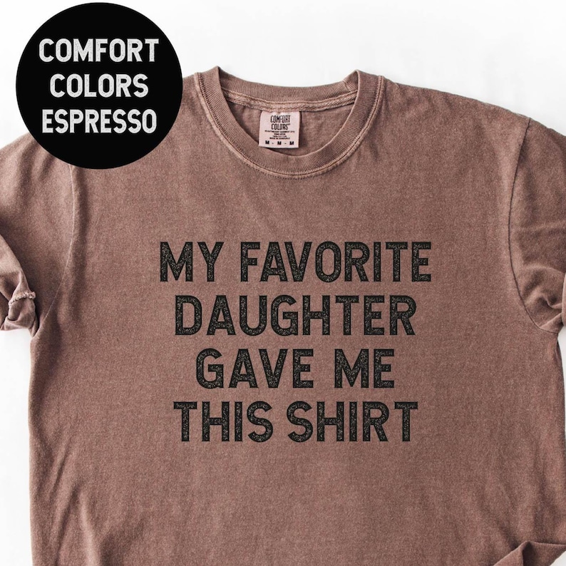 May include: Espresso-colored t-shirt with the text "MY FAVORITE DAUGHTER GAVE ME THIS SHIRT" in black. The shirt has a crew neck and short sleeves. A black circle in the upper left corner reads "COMFORT COLORS ESPRESSO".