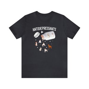 May include: Black t-shirt with the word "ANTIDEPRESSANTS" above an illustration of a pill bottle spilling out cartoon dogs. The dogs are various breeds and colors, with hearts scattered around them.