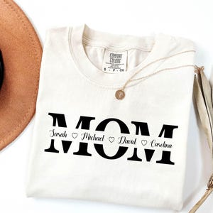 May include: A white t-shirt with the word "MOM" printed in black with a line separating the letters. The names "Sarah", "Michael", "David", and "Carolina" are printed below the letters with a heart symbol between each name.