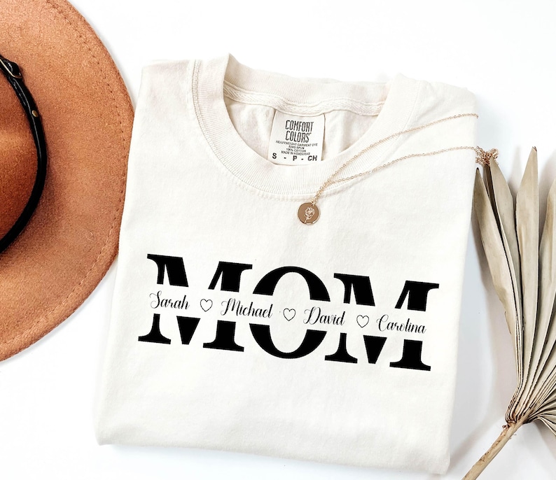 May include: A white t-shirt with the word "MOM" printed in black with a line separating the letters. The names "Sarah", "Michael", "David", and "Carolina" are printed below the letters with a heart symbol between each name.