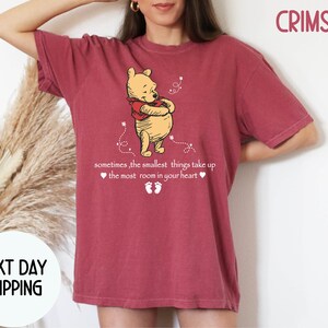 Peut inclure: Un t-shirt rouge fonc&eacute; avec une image de dessin anim&eacute; de Winnie l'ourson et le texte "sometimes, the smallest things take up the most room in your heart".